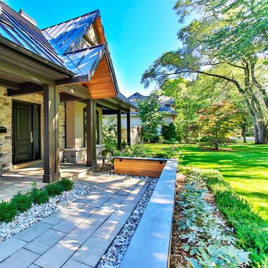 Modern house with stone facade, metal roof, and landscaped garden featuring trees and shrubs. Bright, sunny day enhances the vibrant greenery.