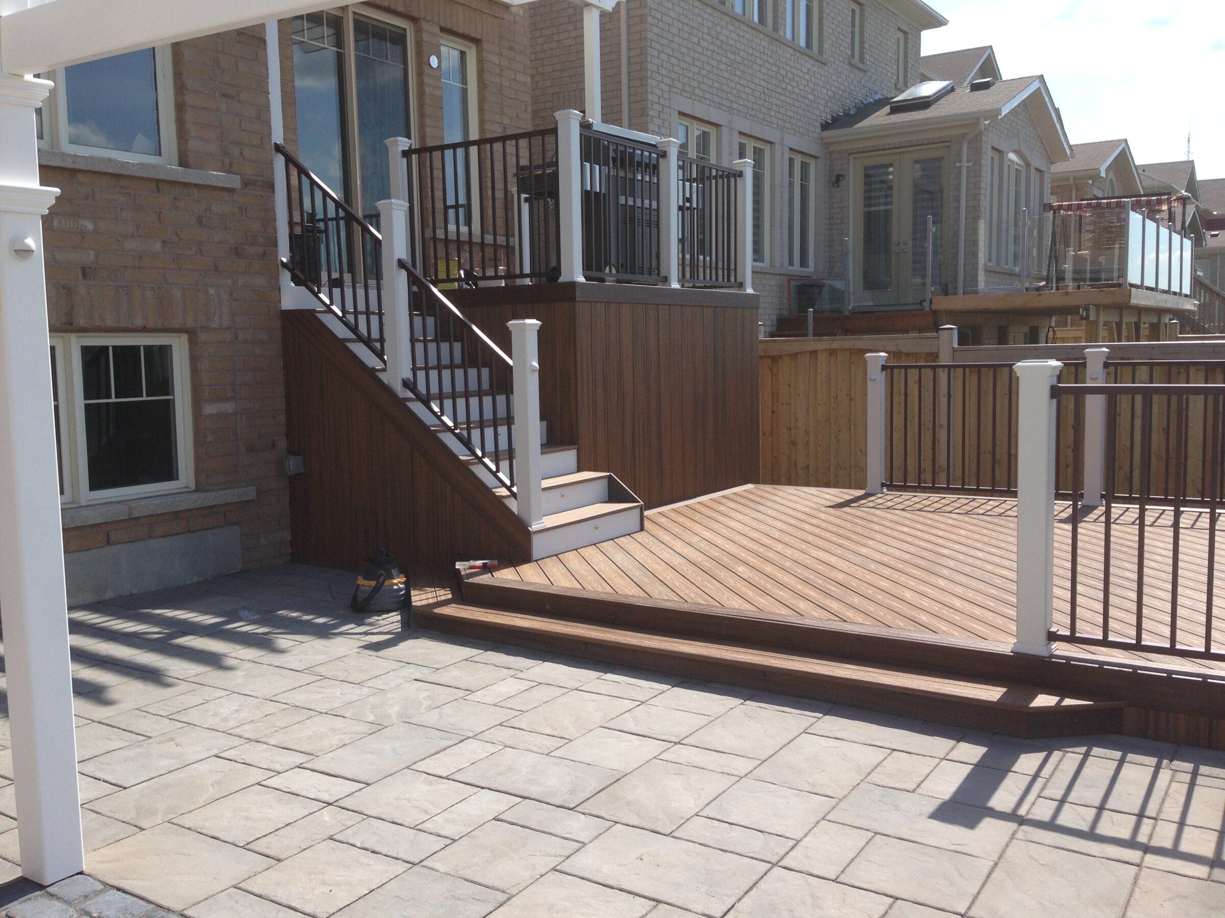 Modern backyard with wooden deck, metal railings, and stone patio; adjacent to brick houses with large windows. Sunny, clear sky.