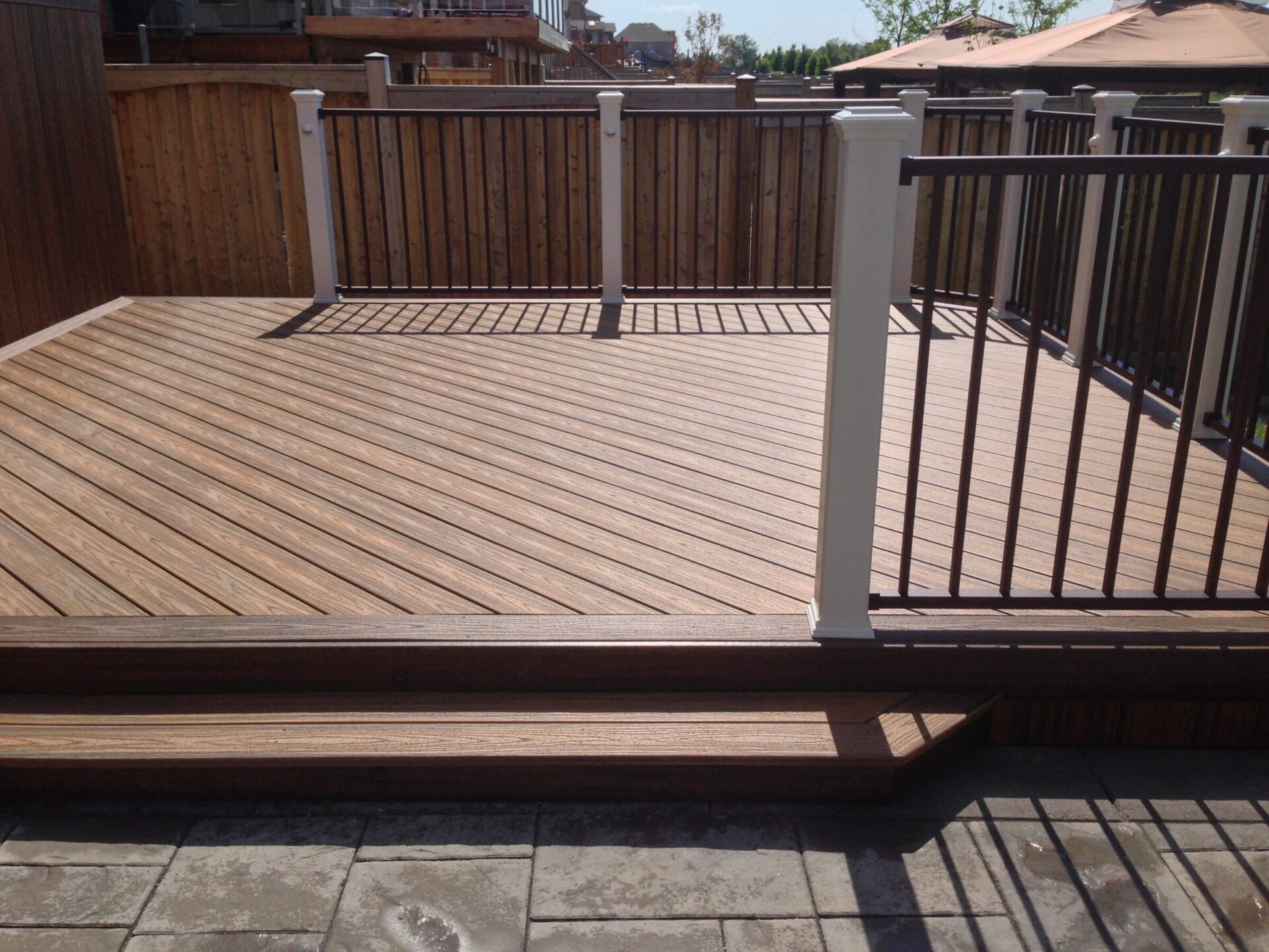 A wooden deck with white and dark railings, surrounded by a wooden fence. Paved area in foreground, with gazebo structures visible.