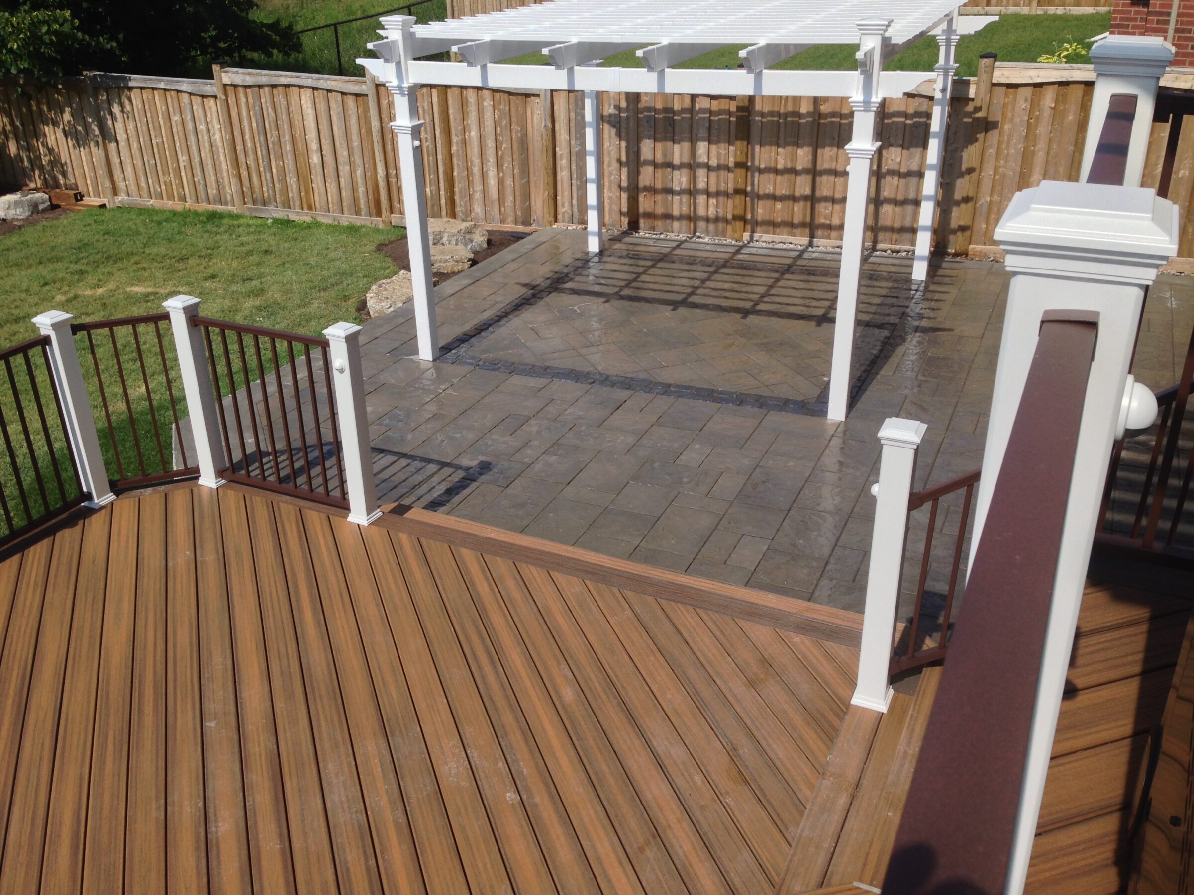 A wooden deck with white railing overlooks a paved patio with a wooden pergola in a fenced backyard on a sunny day.