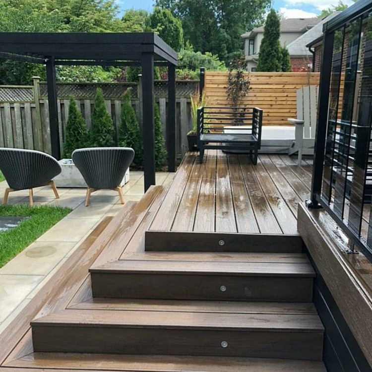 A modern backyard with wooden decking, pergola, and seating area. Lush greenery surrounds, creating a serene outdoor space for relaxation and gathering.