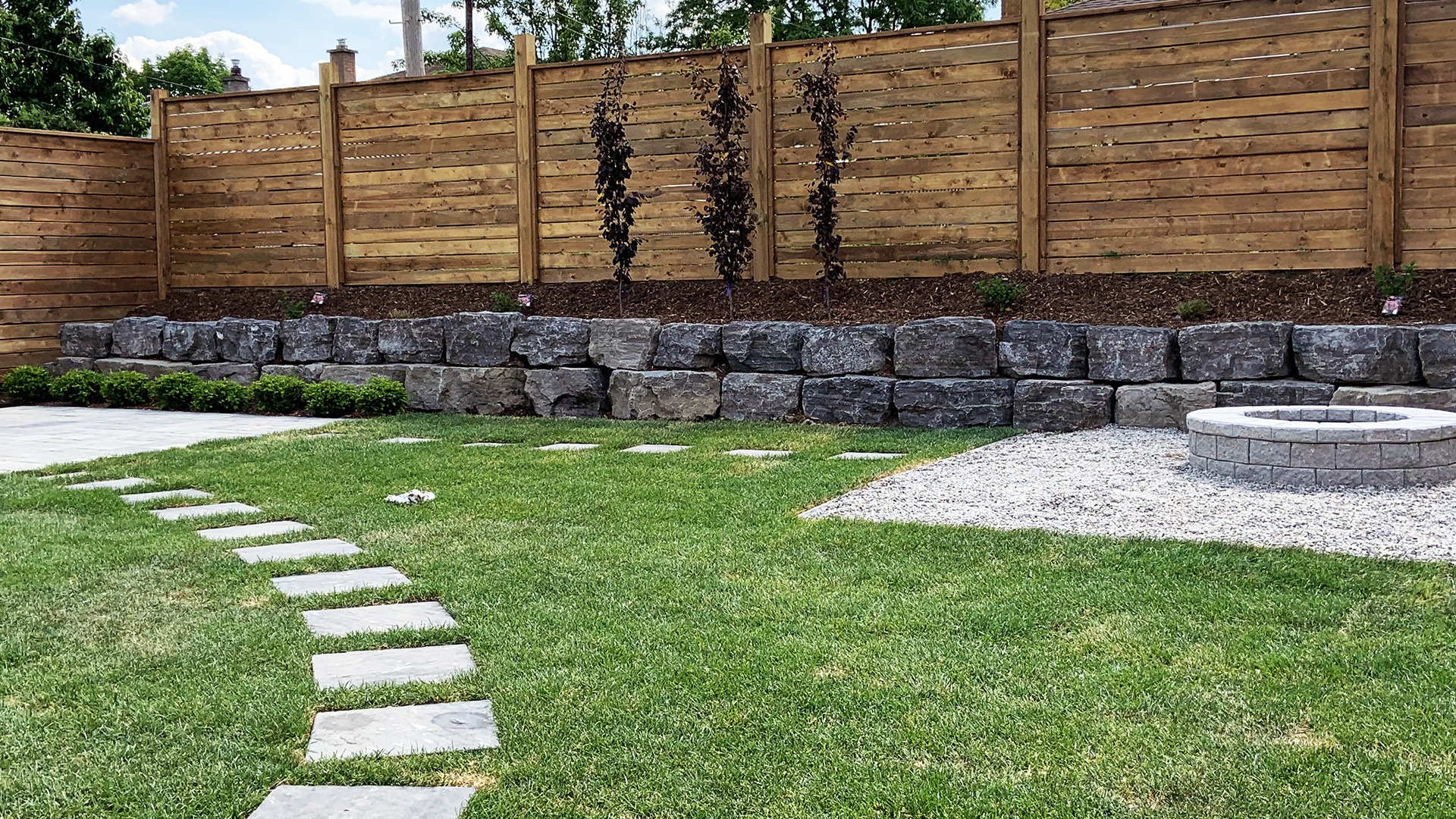 A neatly landscaped backyard features stepping stones, a stone-lined fire pit on gravel, and a wooden privacy fence with climbing plants.