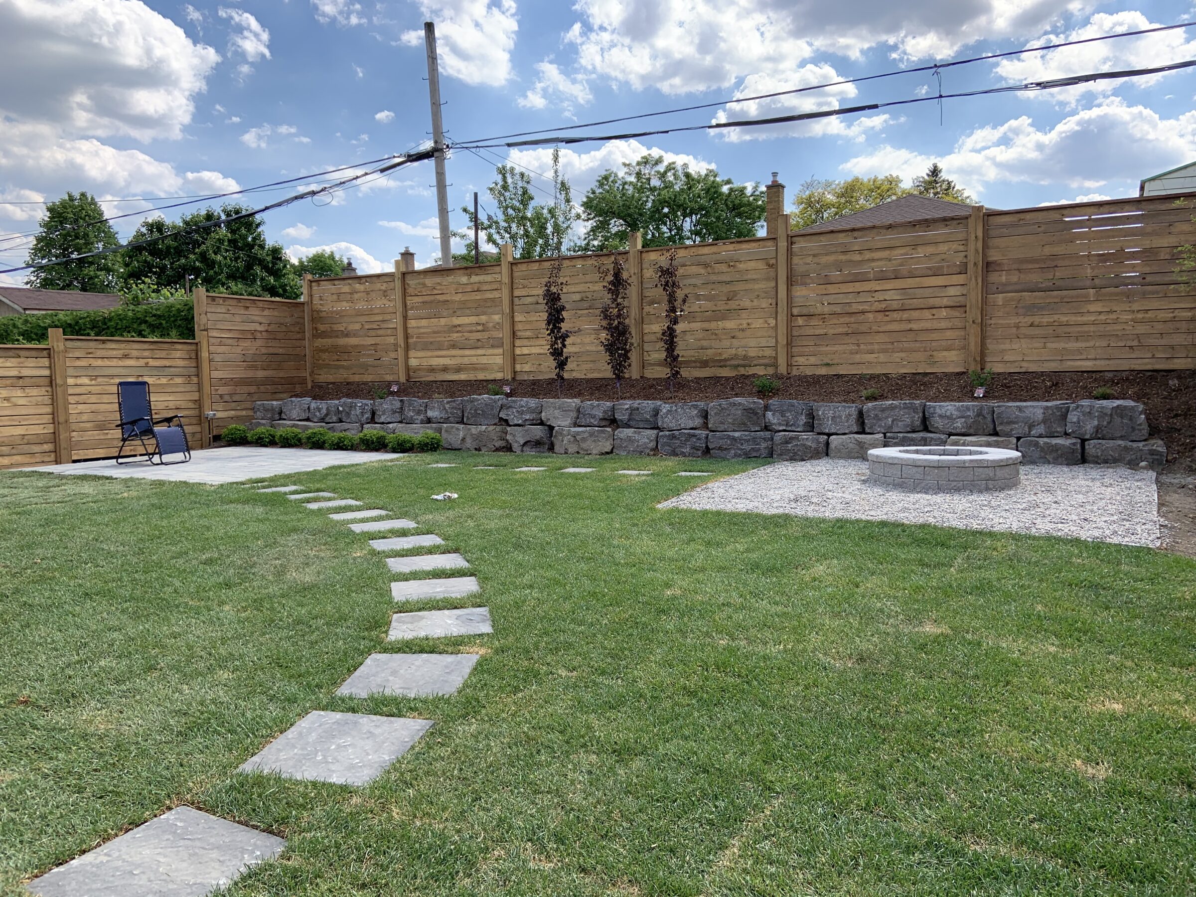 A backyard with a circular stone fire pit, stepping stones, wooden fence, and a blue reclining chair under a partly cloudy sky.