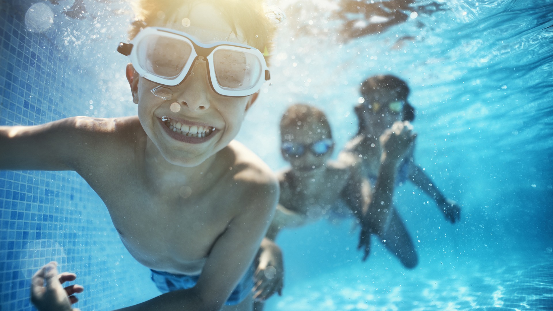 Three children underwater in a swimming pool, wearing goggles, smiling. Bright sunlight filters through the water, creating shimmering patterns.