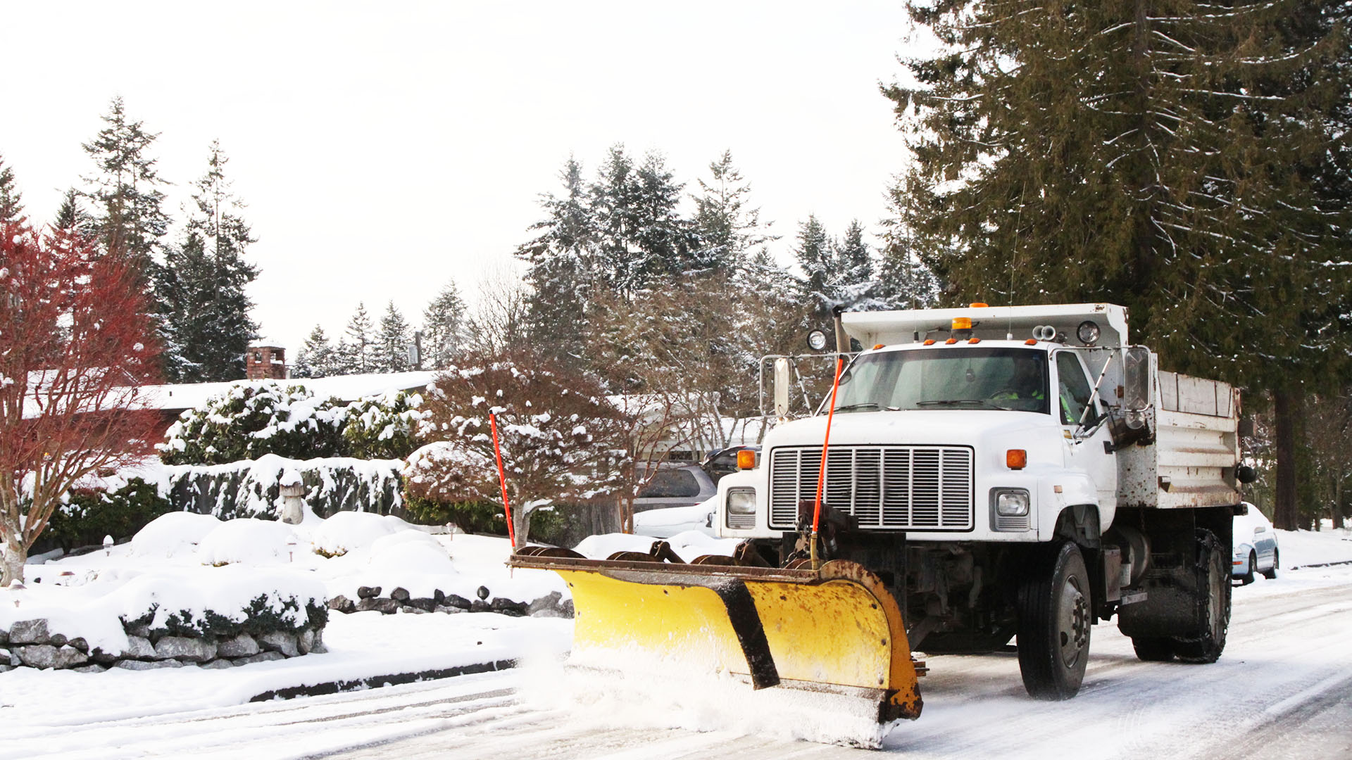 A snowplow truck clears a snow-covered residential street lined with trees and homes during winter, ensuring safe passage for vehicles.