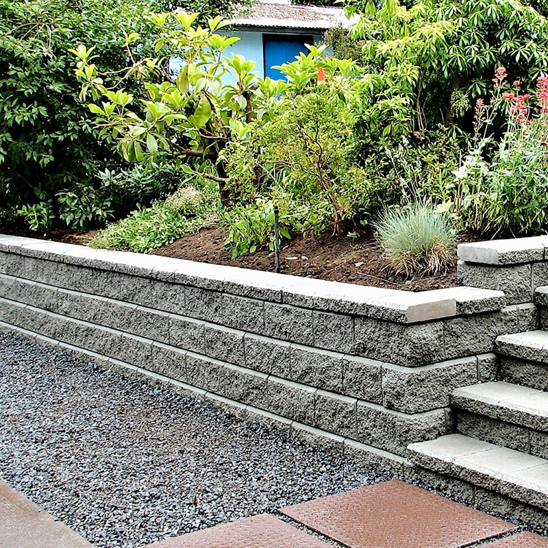 A garden with a stone retaining wall and steps, surrounded by lush green bushes and gravel pathway. No recognizable landmarks visible.
