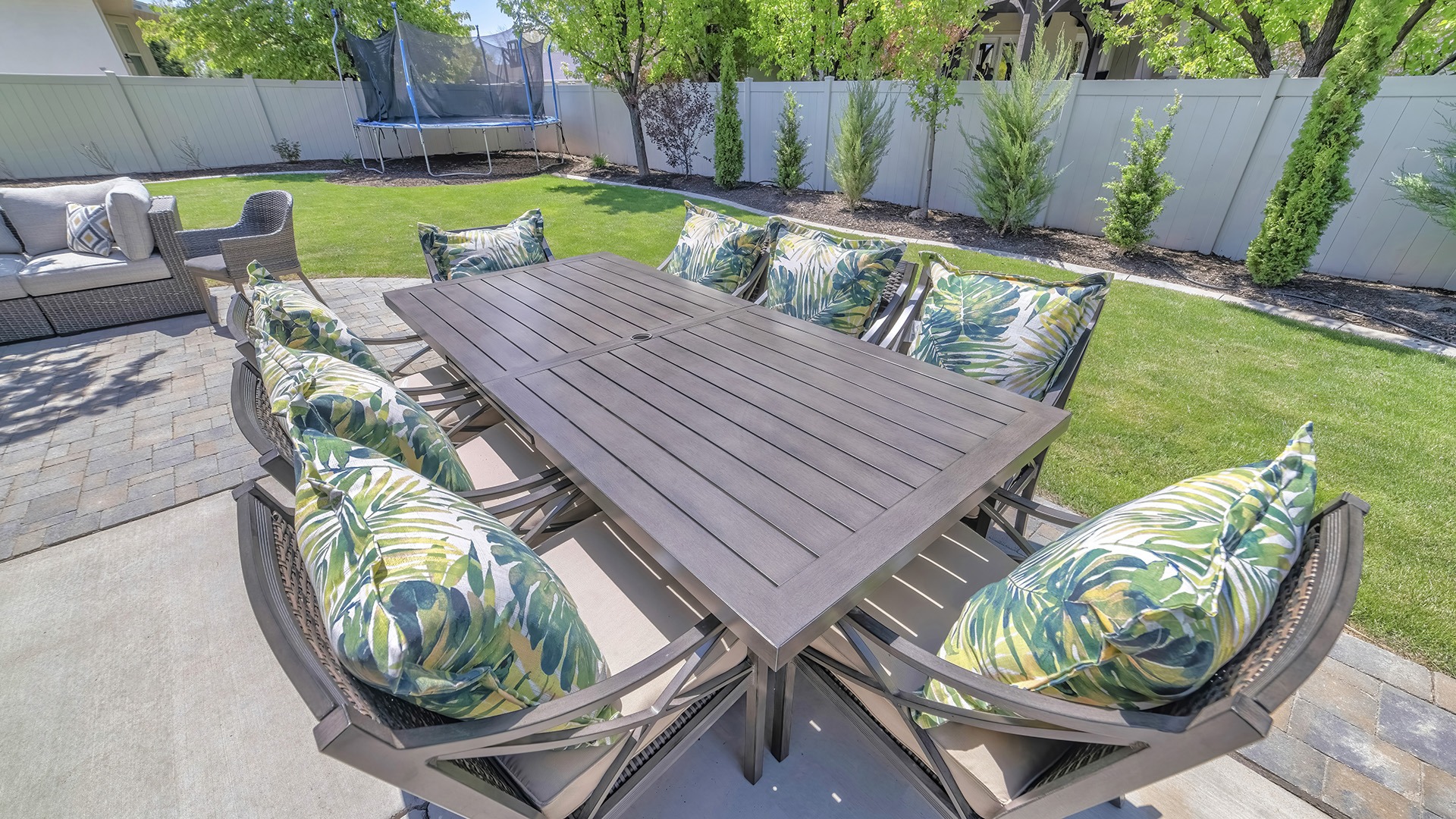 Outdoor patio set with cushioned chairs, wooden table, trampoline, and sofa on paved surface surrounded by green lawn and trees.