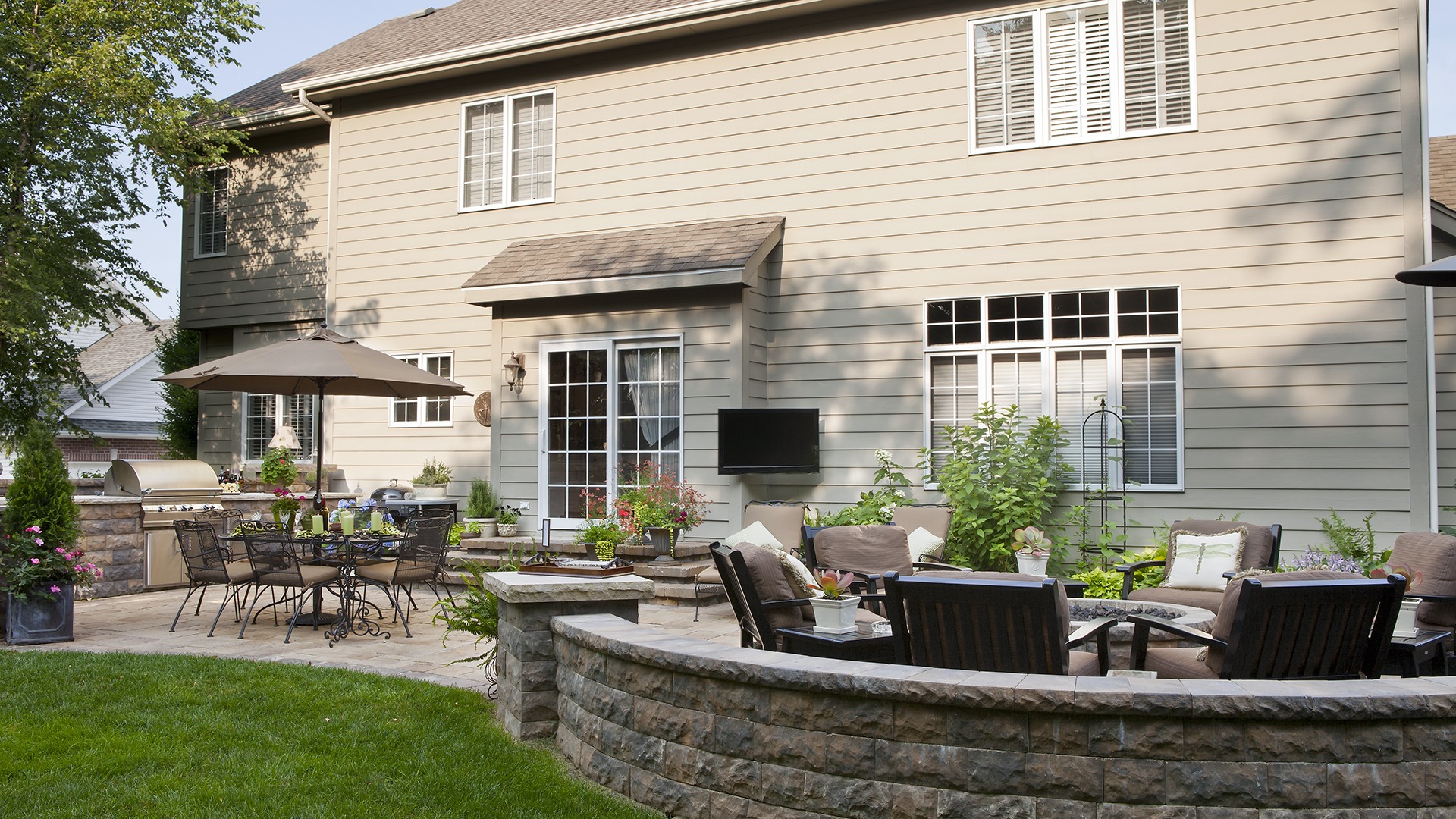 A cozy backyard features patio furniture, a dining table with umbrellas, barbecue grill, and flourishing plants against a two-story house backdrop.