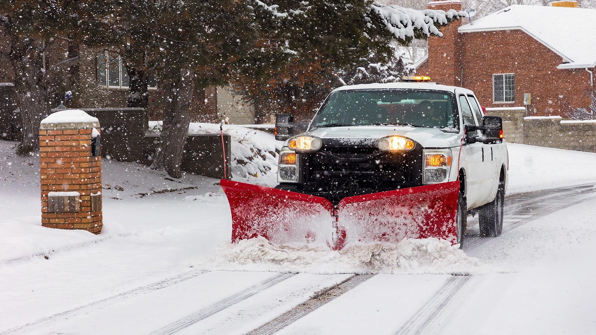 A snowplow truck clears a snow-covered residential street during snowfall, surrounded by brick houses and trees, ensuring safe winter driving conditions.