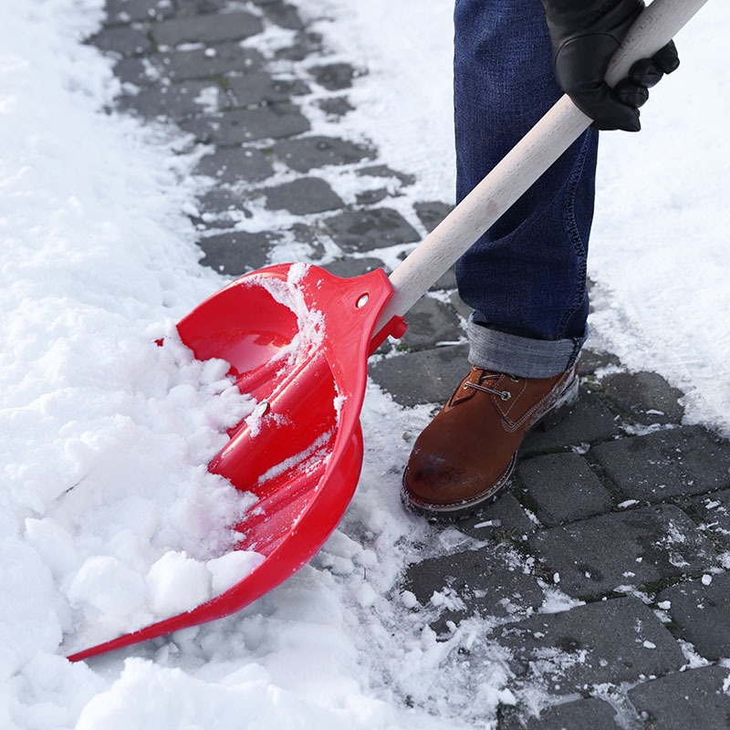 A person in jeans and brown shoes uses a red snow shovel to clear snow from a paved path on a cold day.