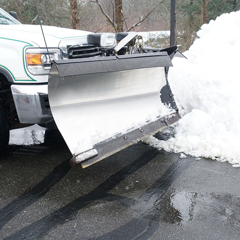 A truck is equipped with a snowplow, clearing snow on an asphalt surface amidst a snowy, wooded backdrop.