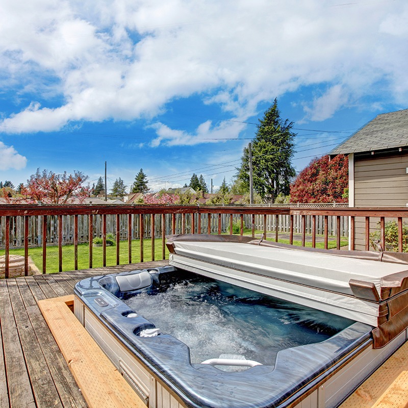 A backyard deck with a bubbling hot tub, surrounded by trees, a wooden fence, and a cloudy blue sky above.