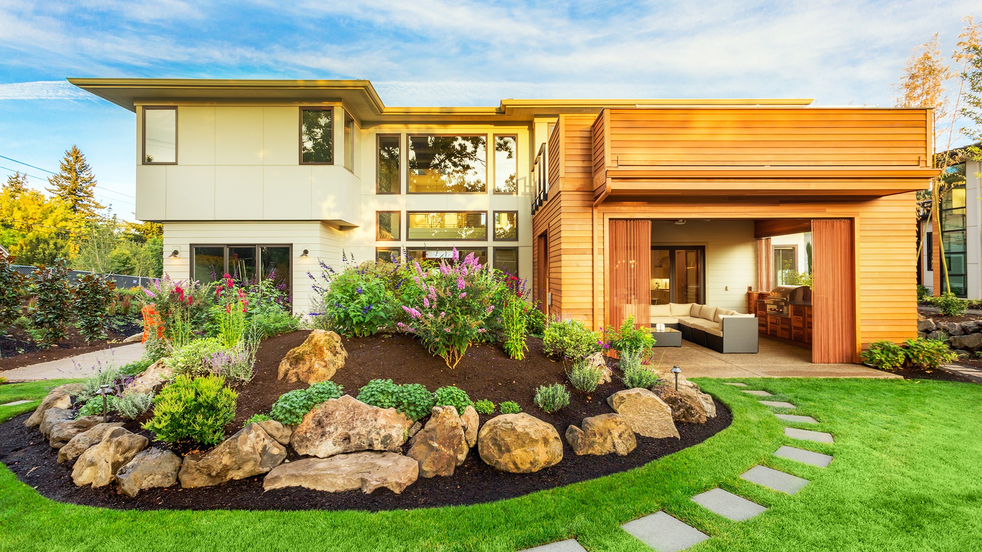 Modern house with large windows, wooden exterior, and lush landscaping. Stone path leads to seating area on patio, surrounded by vibrant garden.
