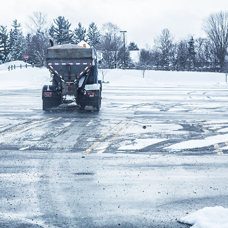 A truck spreads salt on a snowy parking lot, surrounded by bare trees. Snow and ice partially cover the asphalt and surrounding areas.