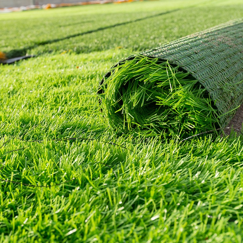 A roll of vibrant green artificial grass lies on a smooth lawn, ready for installation, under a clear, sunlit sky.