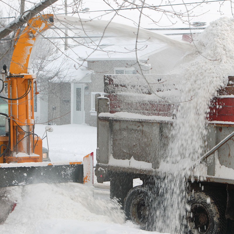 A snowblower transfers snow from the street into a large truck, with houses visible in the snowy background. No recognizable landmarks present.