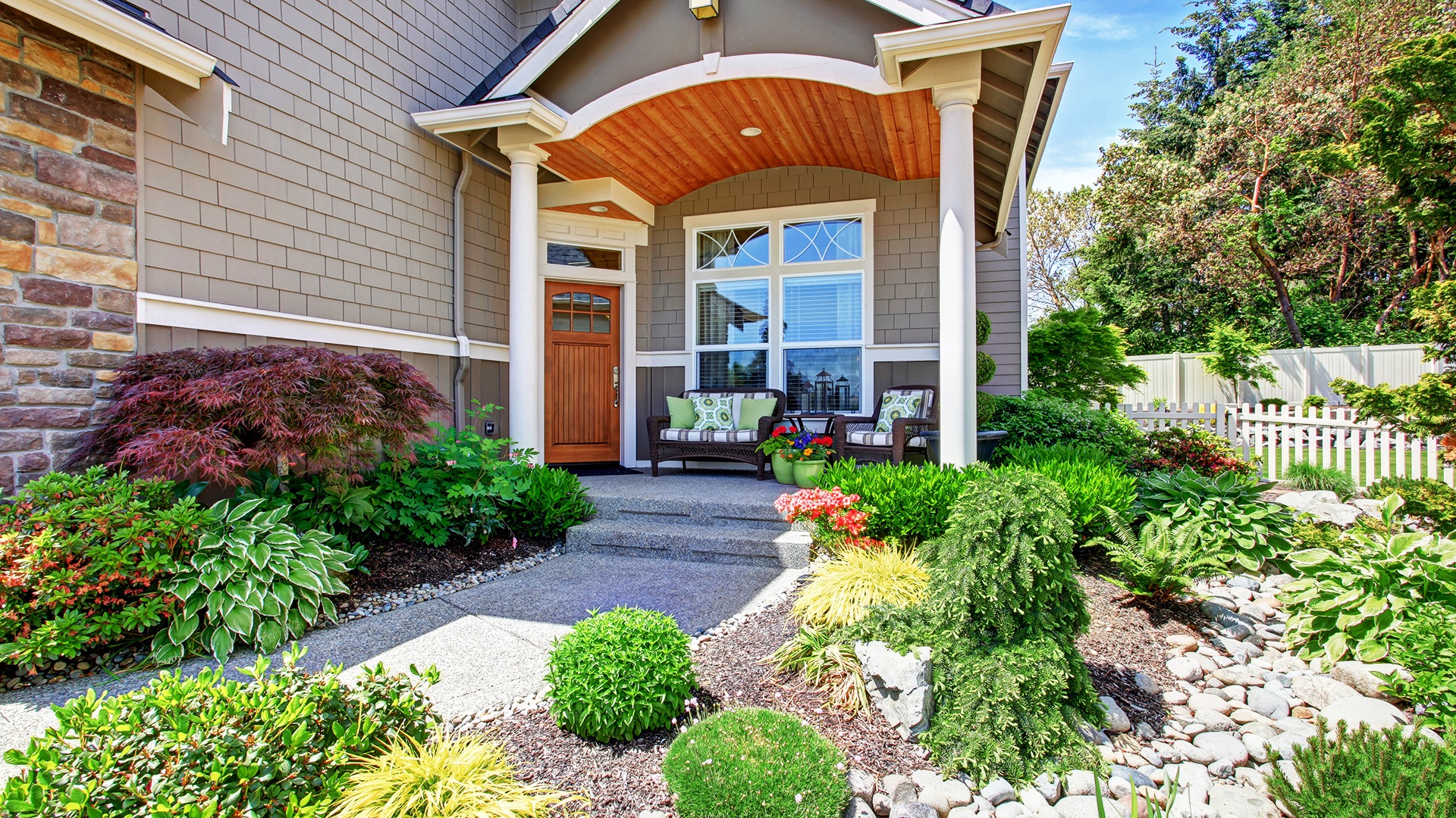 A charming house entrance features a cozy porch, seating area with cushions, and a beautifully landscaped garden with lush greenery and colorful flowers.