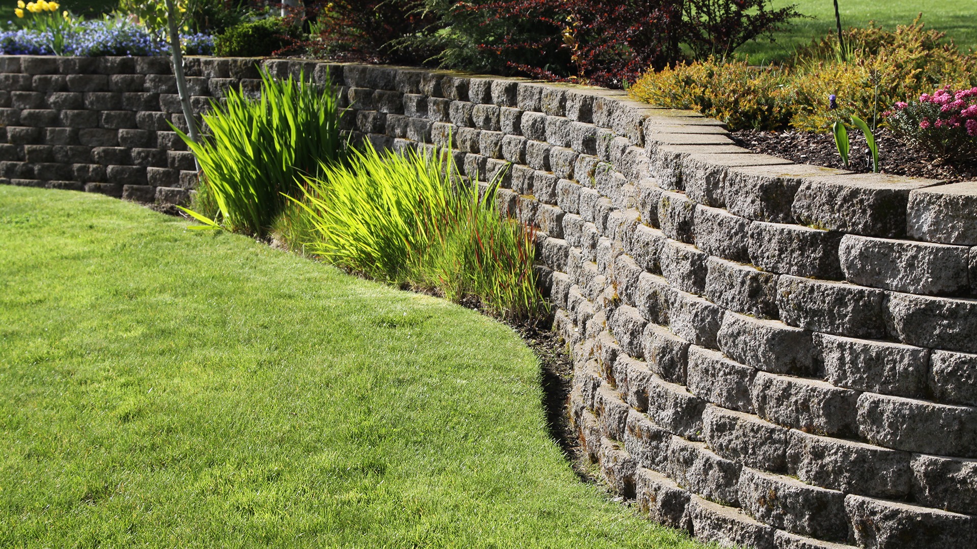 Curved retaining wall with stacked stones, vibrant green plants, colorful flowers, and well-maintained grassy area in a sunny outdoor garden setting.