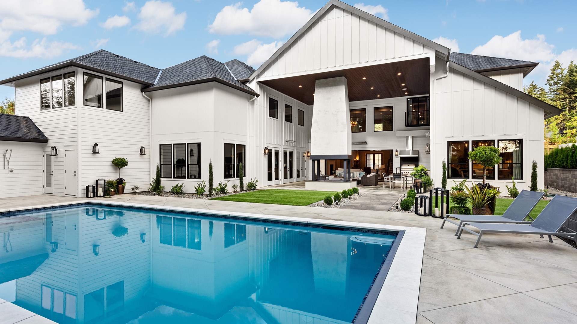 Modern white house with large windows and a spacious backyard featuring a pool, lounge chairs, and landscaped garden under a partly cloudy sky.