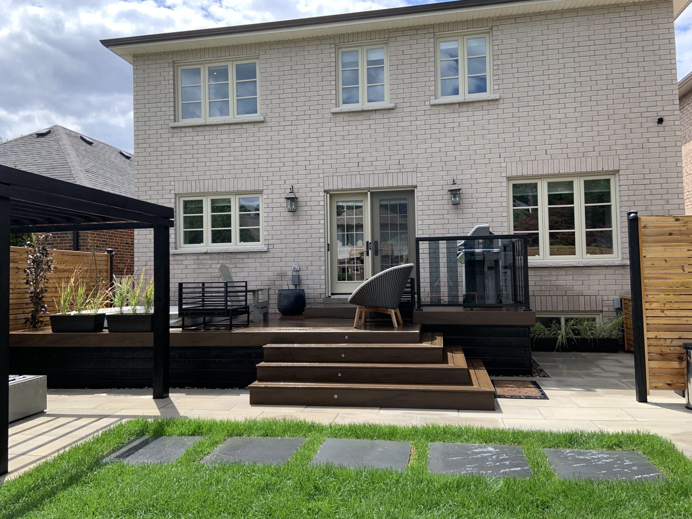 Two-story brick house with a wooden deck, seating area, and pergola. Lush green lawn and stepping stones in the foreground.