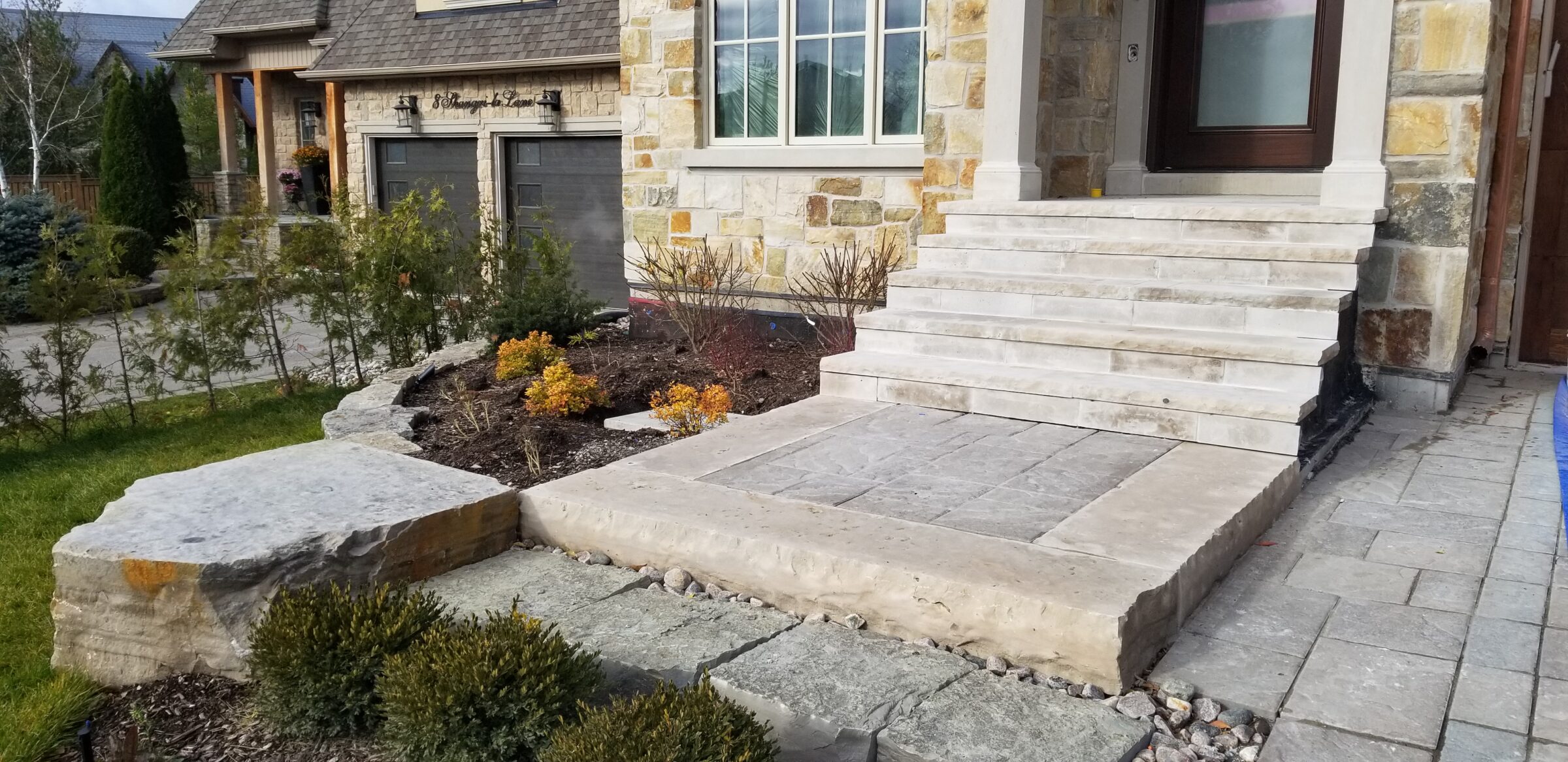 A stone house with a front walkway, steps, and landscaped garden featuring shrubs. Two garage doors are visible in the background.