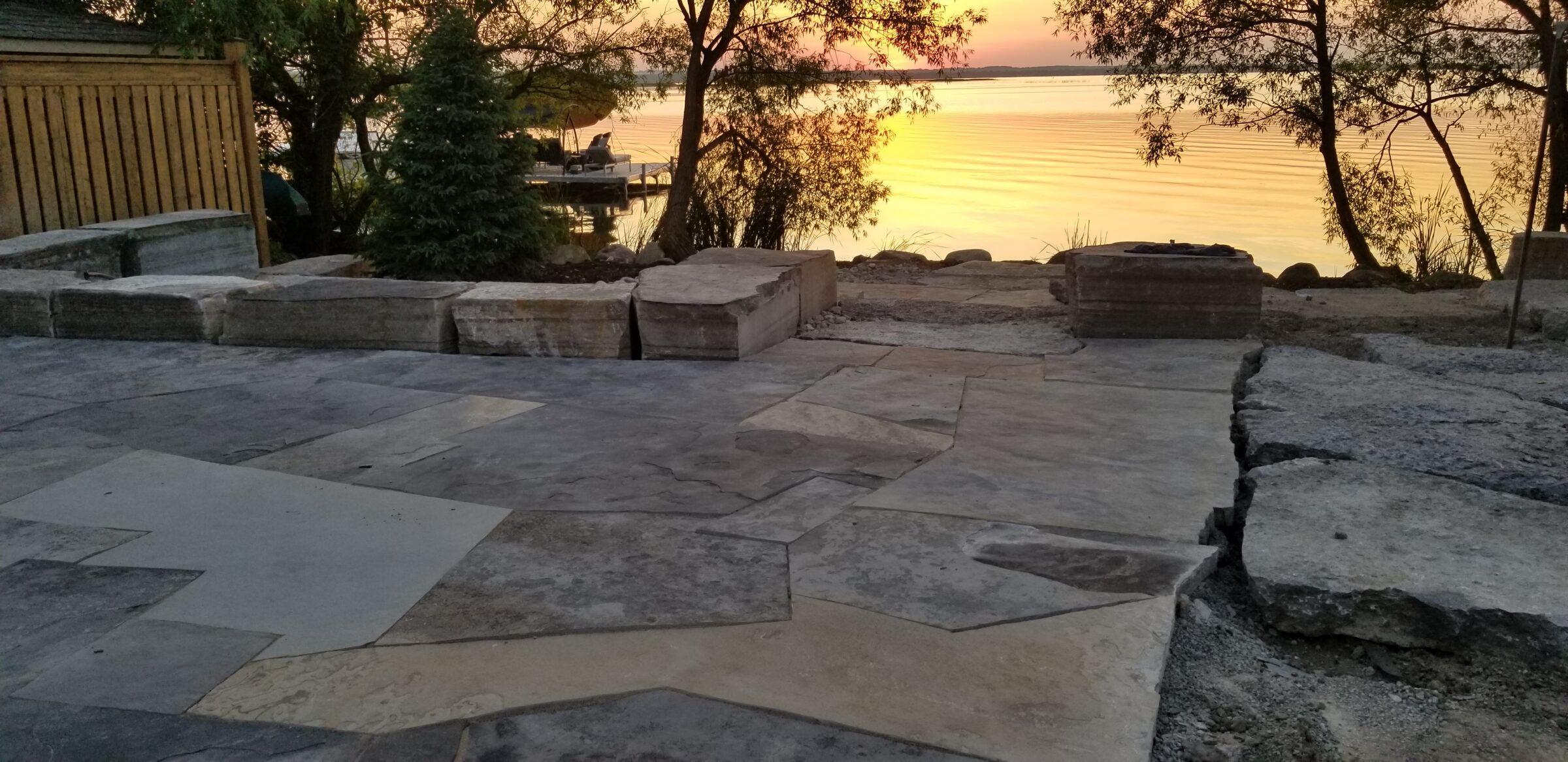Patio ideas using a stone patio overlooking a serene lake at sunset; trees and a small dock are visible.