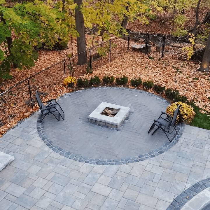 A circular stone patio with a central fire pit, surrounded by trees with fall foliage and two chairs.