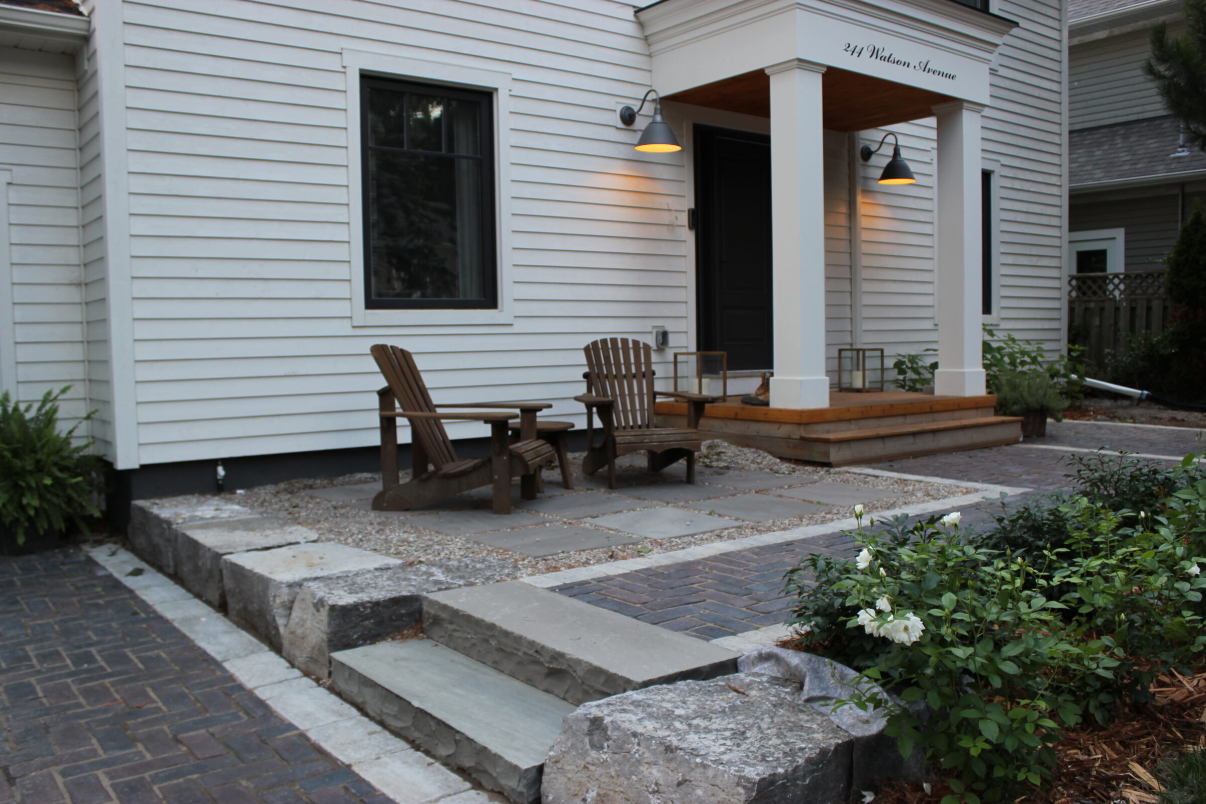 A modern white house features a wooden porch and seating area, surrounded by stone steps, plants, and elegant outdoor lighting.